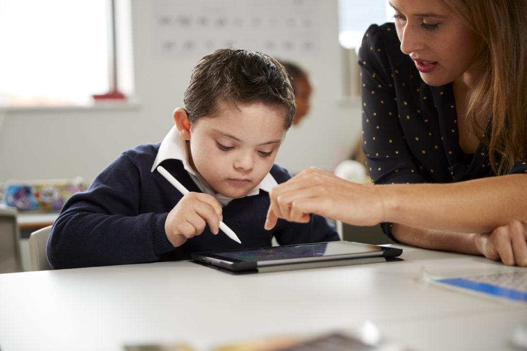 在一所小学的教室里，年轻的女老师正在和一个患有唐氏综合症的男孩一起使用平板电脑, front view, close up