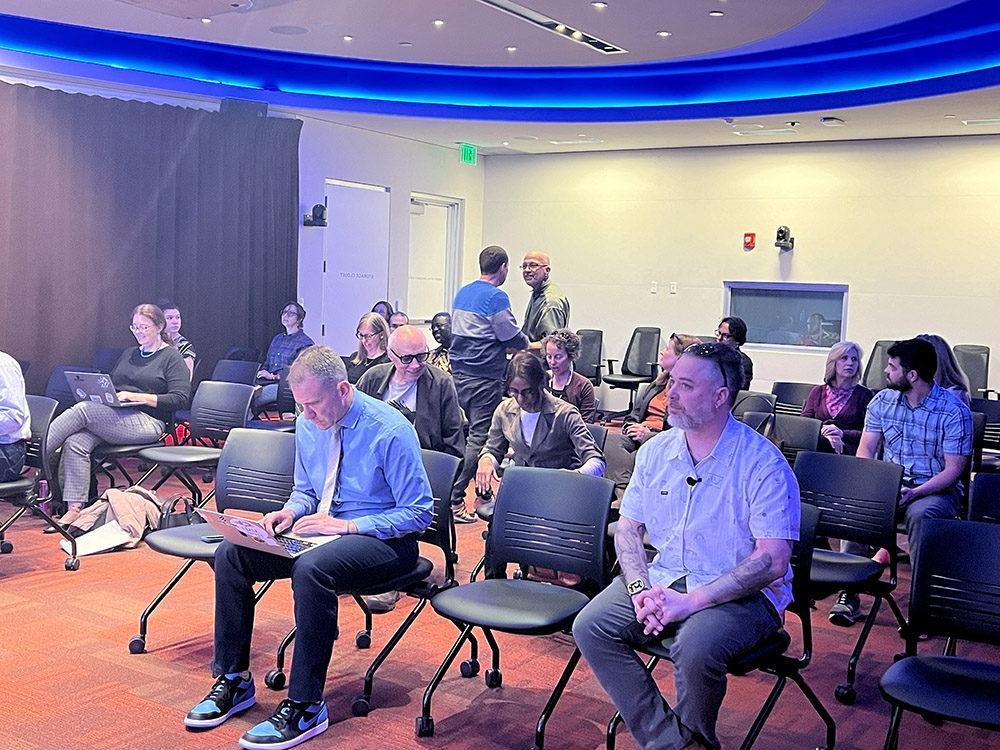 这张图片显示了特拉维斯·马辛格尔在大学课堂上使用人工智能的演讲, 由丹佛大都会州立大学英语系主办并在CAVEA举行的活动. 与会者, 谁是主要的教员, 坐在一排椅子上, 有些人在笔记本或笔记本上做笔记. 房间有一个弯曲的布局，沿着天花板的边缘有蓝色LED照明.